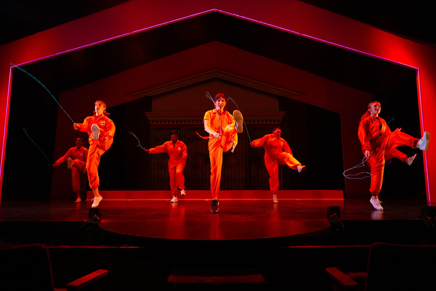 Five performers in orange prison jumpsuits jump rope in formation on a red-lit stage.
