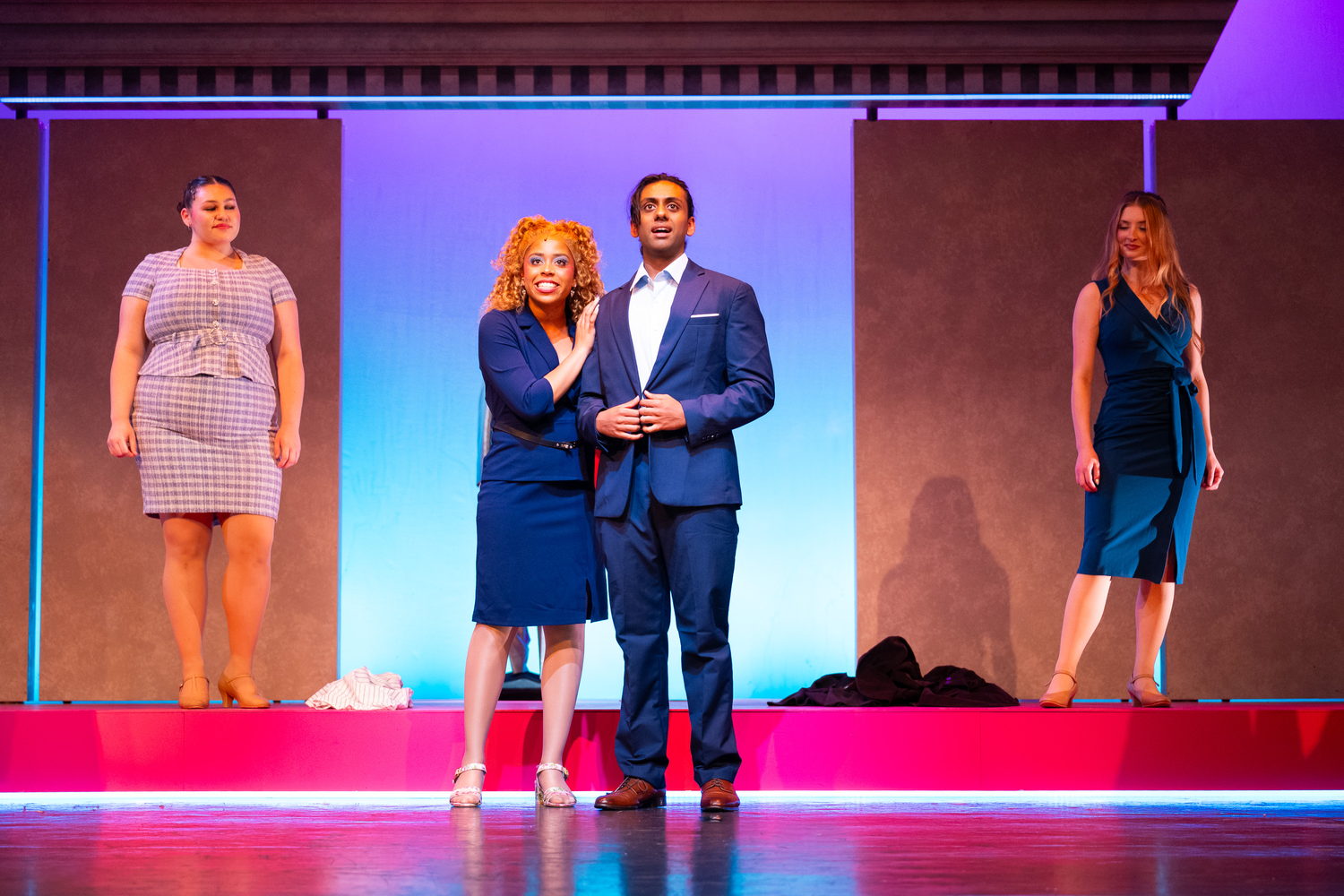 Elle Woods and Emmett stand arm in arm in business attire on a courthouse set, with two other cast members looking on.