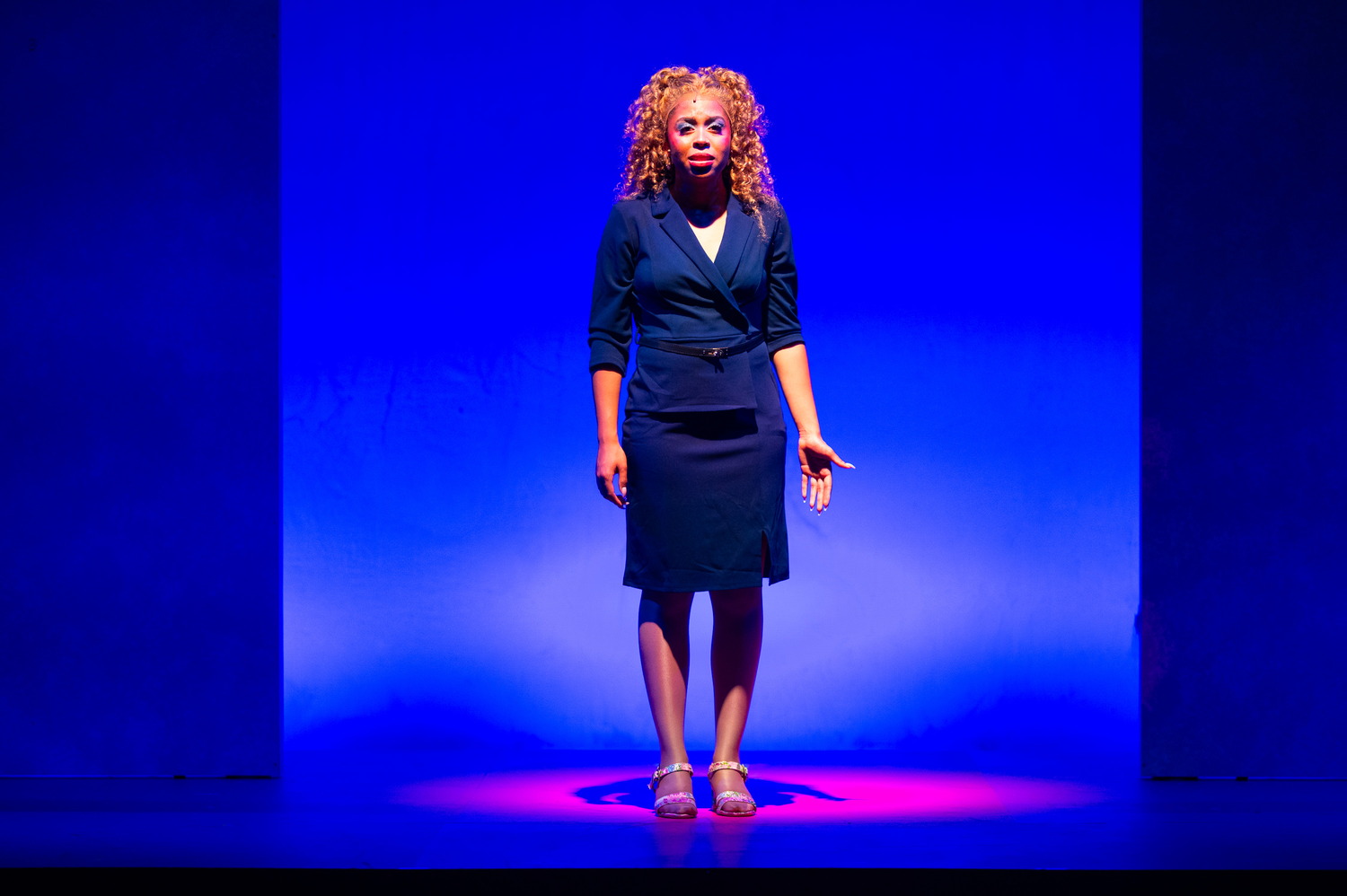 Elle Woods stands alone in a navy suit dress in a pool of blue light on a bare stage.